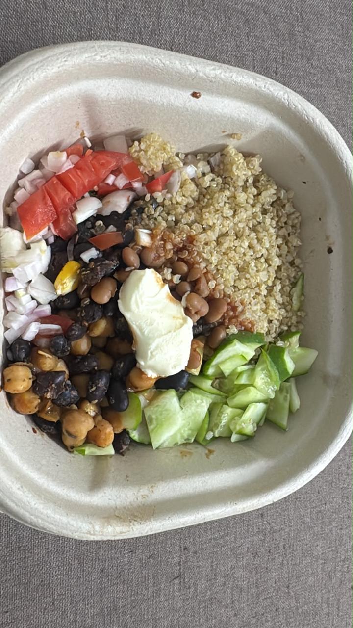 A takeaway bowl with quinoa, black beans, chickpeas, chopped tomato, onion, cucumber and a spoon of curd