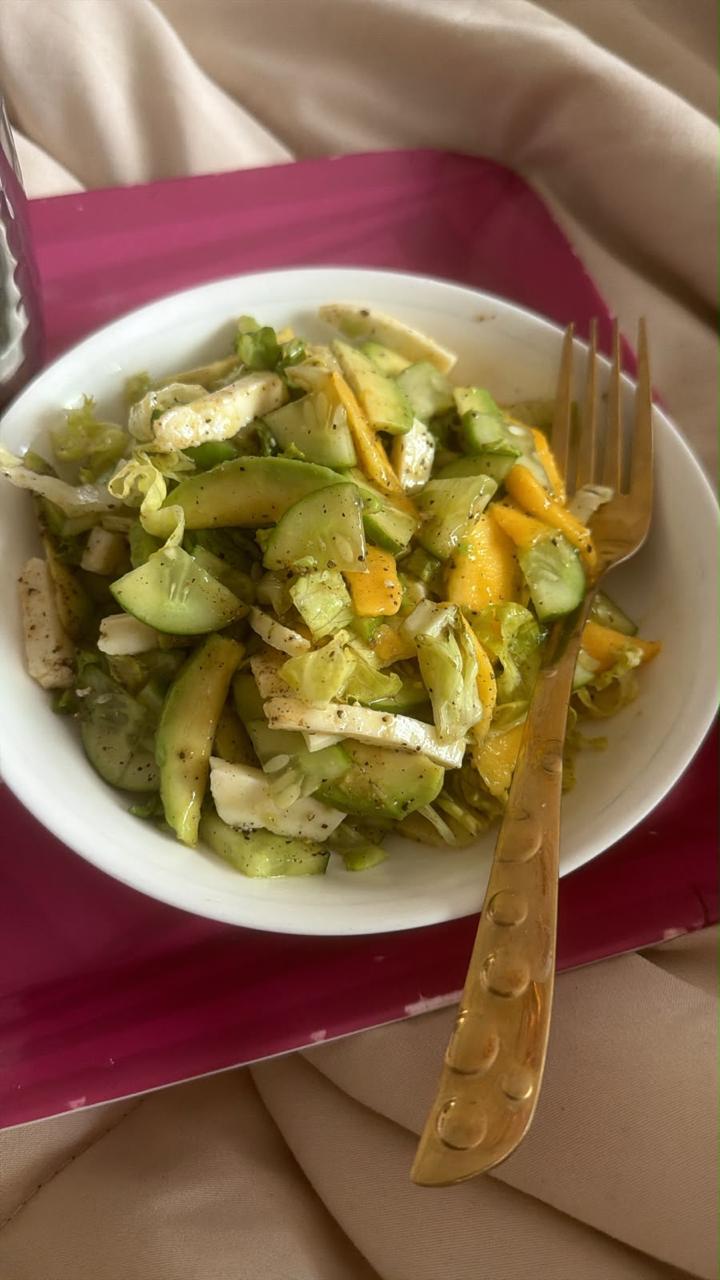 A white bowl of salad with avocado, cucumber, mango and crisp lettuce, with a gold fork on a magenta tray