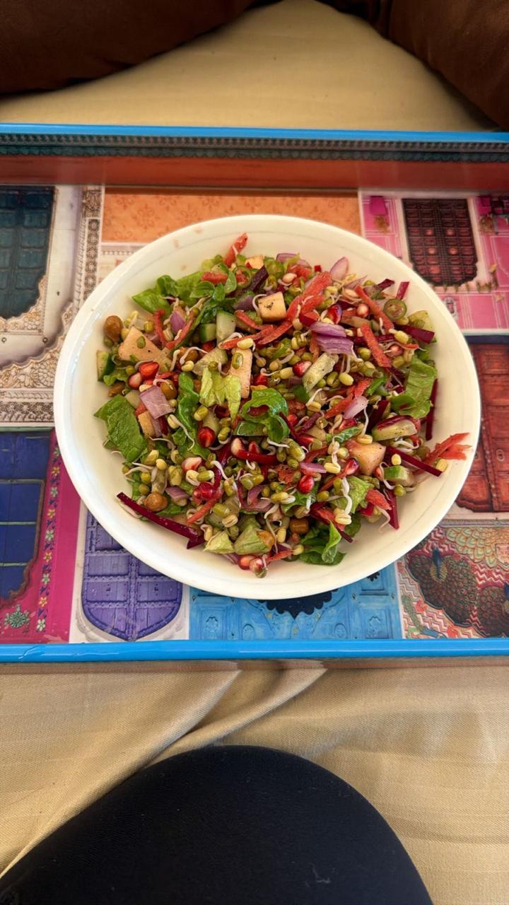 A white bowl of sprouted moong salad with pomegranate seeds, cucumber, onion and leafy greens on a printed tray