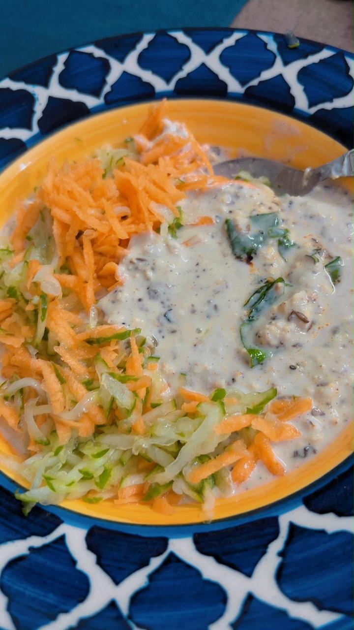 An orange bowl of creamy oats porridge with herbs beside a mound of shredded carrot and cucumber