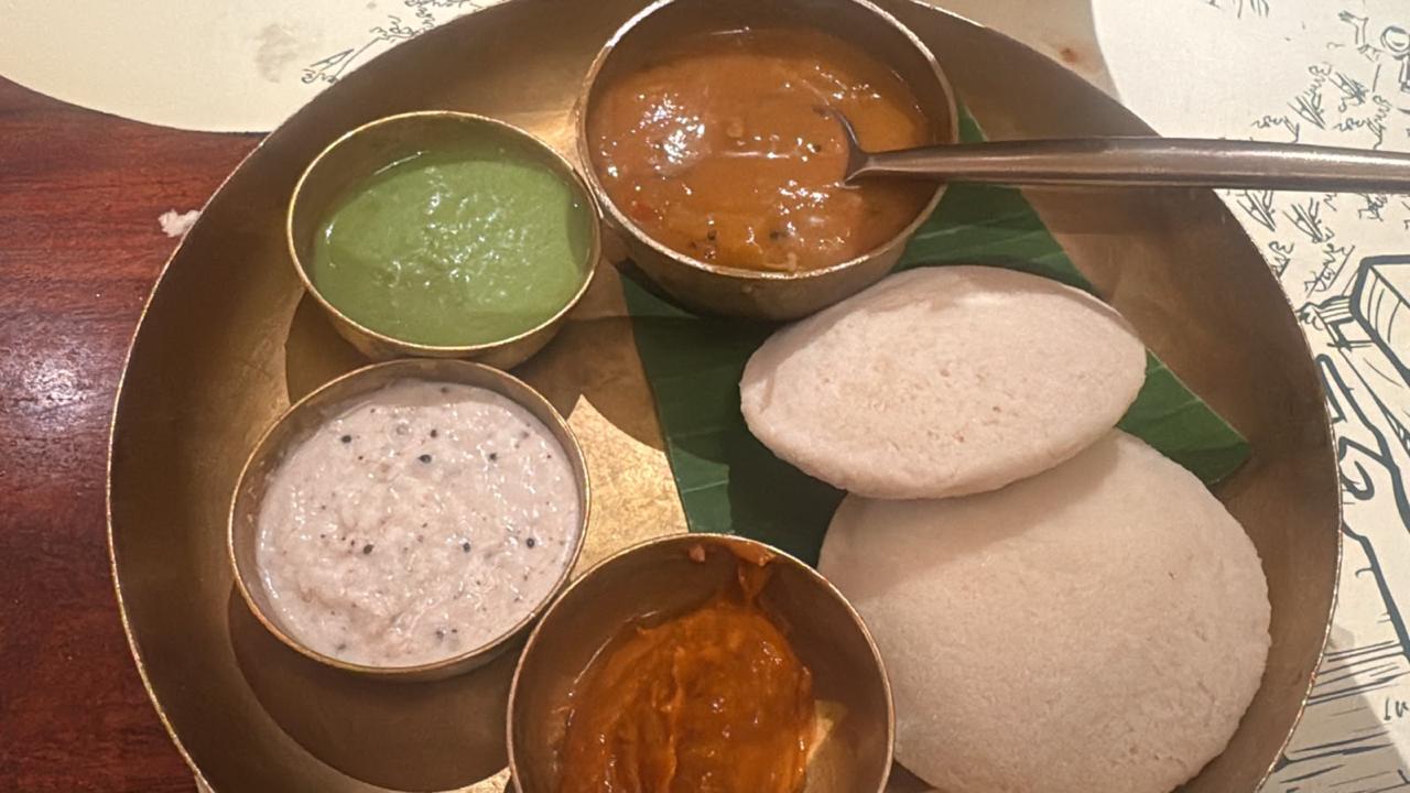 A brass platter with two idlis, sambar, a white chutney, a green chutney and a red chutney