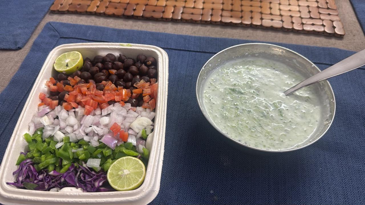 A takeaway box of chopped tomatoes, onion, capsicum, purple cabbage, black chickpeas and lime wedges beside a bowl of cucumber raita