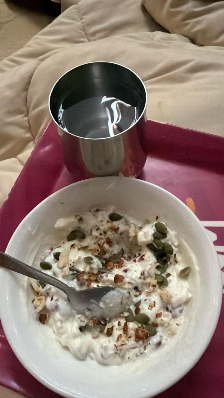 A white bowl of curd topped with pumpkin seeds and chopped nuts, beside a steel glass of water on a pink tray