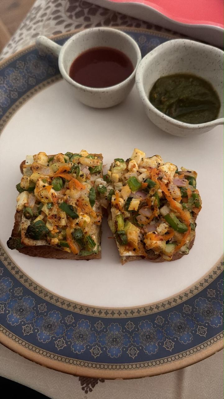 Two slices of open-faced toast topped with paneer and vegetables, with two small cups of chutney