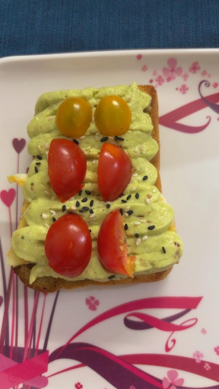 A slice of toast with avocado spread, cherry tomato halves and black sesame seeds on a floral plate