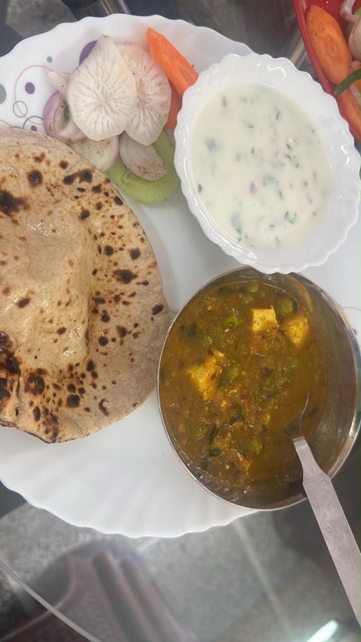 A tandoori roti beside a steel bowl of paneer and pea curry, a bowl of curd and sliced onion